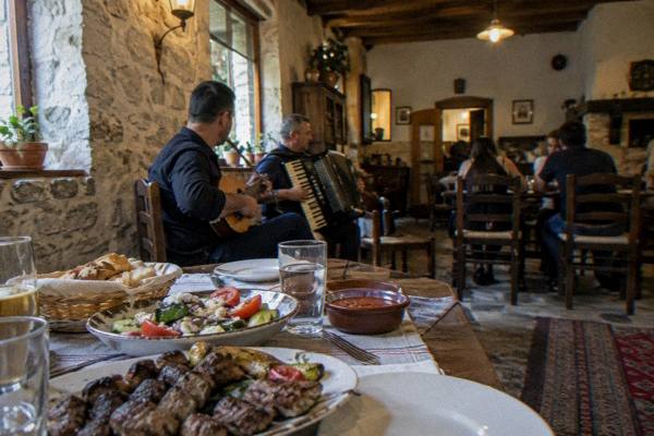 A restaurant table laid with grilled meat, salad and bread beside a small band with a lute and an accordion in a rustic dining room, Blloku