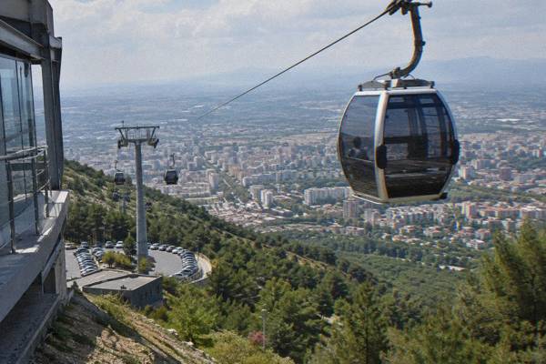 A large gondola cabin rising on a steel cable over pine and scrub slopes above the city, hazy plain far below, Dajti Ekspres lower station