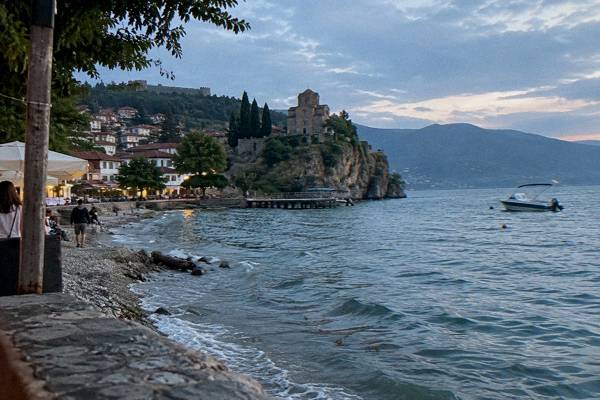 A small hilltop stone church on a cliff above a large clear lake with wooded hills and a pebble shore, Ohrid