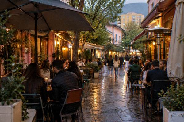 A busy narrow street of bar terraces at night with neon-free warm lights and groups at tables, seen from the end of the lane, Blloku