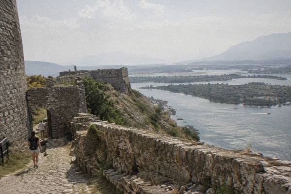 Weathered stone castle walls on a rocky hill above a wide flat lake plain with distant mountains, Rozafa Castle