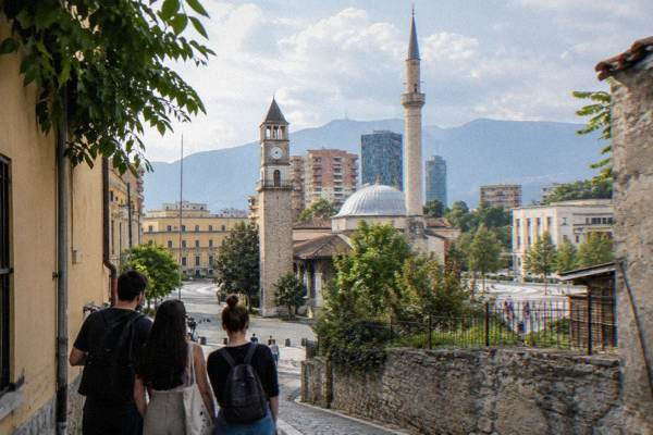 A small group on a quiet side street behind the square looking at a tall stone clock tower and a domed mosque in warm daylight, seen from behind, Skanderbeg Square