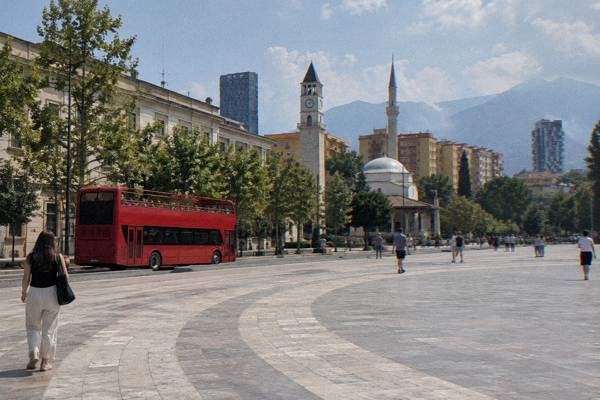 A red open-top double-decker bus without any text stopped beside a wide boulevard with plane trees and a ministry building behind, Skanderbeg Square