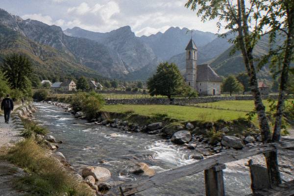 A small stone church tower in a green alpine valley ringed by jagged grey peaks, a river with pale boulders beside it, Theth