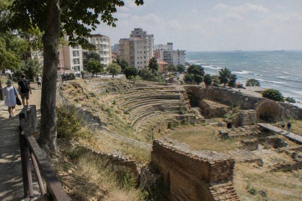 The curved stone tiers of a ruined Roman amphitheatre among modern apartment blocks, with the Adriatic beyond, Durres Amphitheatre