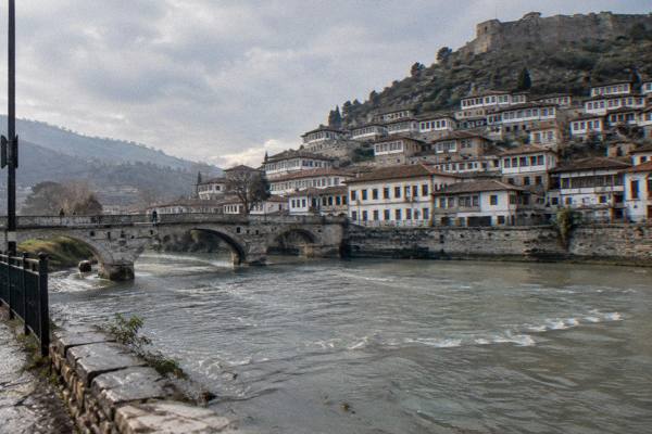 White Ottoman houses with rows of windows stacked up a steep hillside above a river, seen from the far bank in soft daylight, Berat