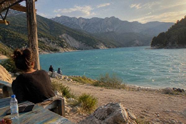 A calm turquoise mountain reservoir with a ridge of bare limestone peaks behind and a gravel viewpoint terrace in the foreground, Lake Bovilla