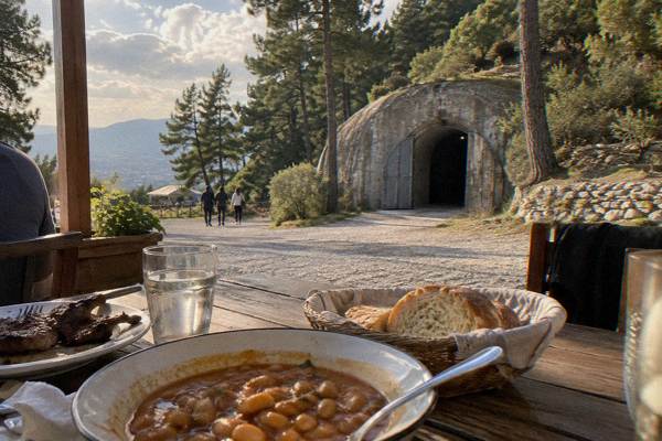 A lunch table with bowls of stewed beans, bread and grilled meat on a terrace at the foot of a pine-covered mountain, Bunk'Art 1