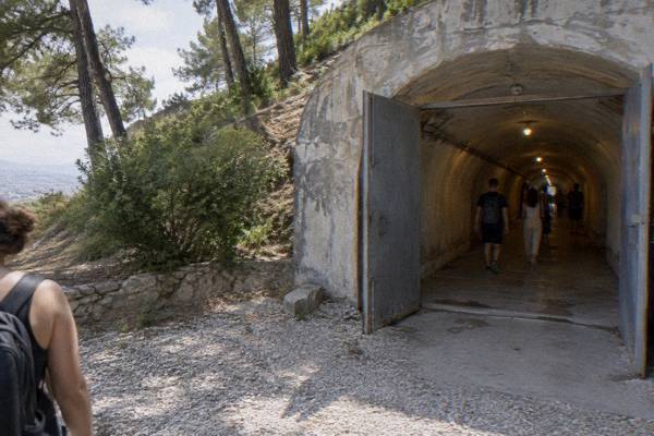 A long underground concrete tunnel corridor with a curved ceiling lit by hanging bulbs and a few visitors walking away, Bunk'Art 1