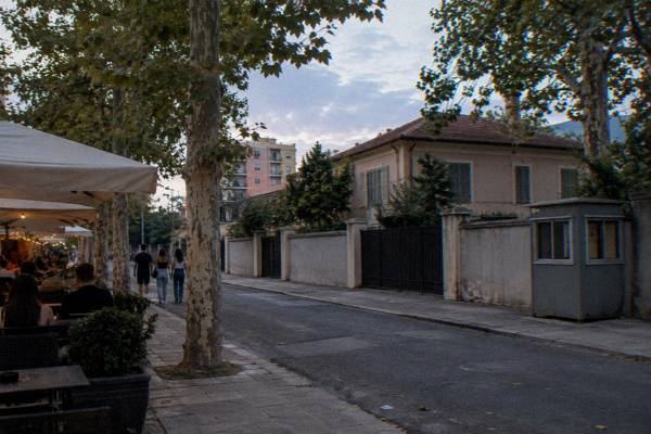 A low villa behind a high wall in a residential street with plane trees, shuttered windows and a small guard post, Blloku
