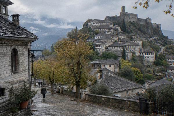 Grey stone tower-houses with heavy slate roofs stacked on a steep hillside under a castle, mountains behind, Gjirokaster