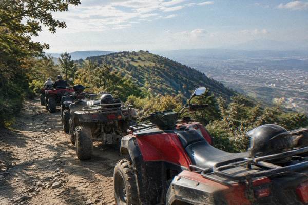 A line of quad bikes parked on a dusty trail among scrub and oak woods with a ridge behind, Mount Dajti