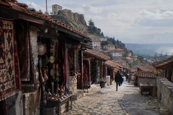 A covered stone bazaar street of small craft shops with carpets and copperware hanging at the doorways below a fortress wall, Kruja