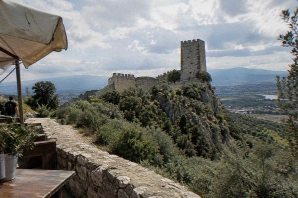 A small stone hilltop castle with a square tower and crenellated wall above olive-covered slopes, Petrela Castle