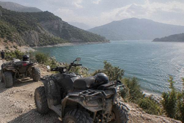 Two quad bikes parked on a dirt track above a blue reservoir with dry hills beyond, helmets resting on the seats, Lake Bovilla