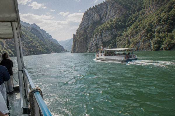 A plain passenger ferry crossing a long narrow green reservoir between steep forested mountain walls with a few visitors on deck, Lake Komani