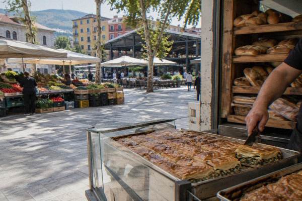 A bakery counter with trays of golden byrek pastry and a hand slicing one, shelves of bread behind, New Bazaar