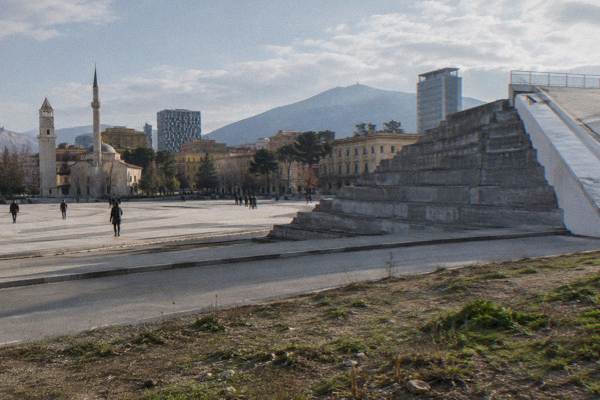 A grey concrete stepped pyramid with a white-clad ramp beside a road, scrubby ground in front, seen from a low angle, Skanderbeg Square
