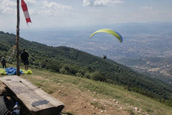 A bright paraglider canopy open in the air over a green mountain slope with the city basin hazy below, Mount Dajti