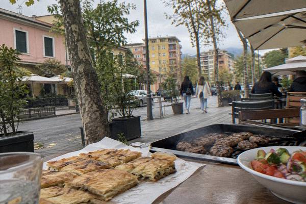 A thin layered byrek pie cut into squares on paper beside a grill tray of qofte and a small salad, Blloku