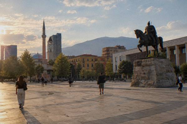 A wide paved square with a bronze a bronze rider on a tall stone plinth in a wide square, a modern museum facade on one side and plane trees, a few visitors crossing, Skanderbeg Square