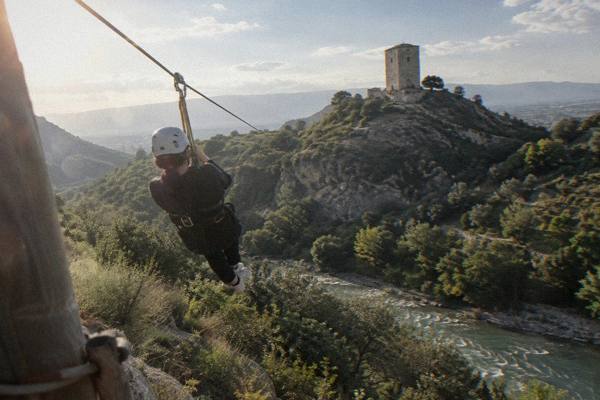 A rider in a harness and helmet seen from behind sliding on a steel cable across a green valley under a clear sky, Petrela Castle
