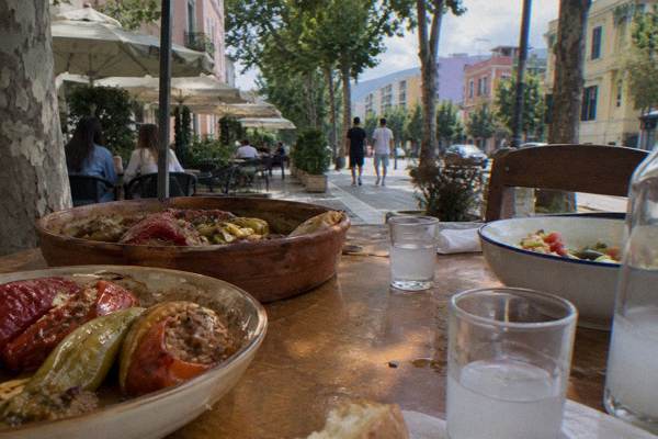 A kitchen table set with bowls of stuffed peppers, a clay baking dish and small glasses of fruit raki, Blloku