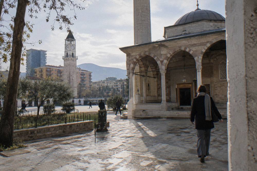 A visitor in long trousers and a scarf entering a historic mosque courtyard in Tirana
