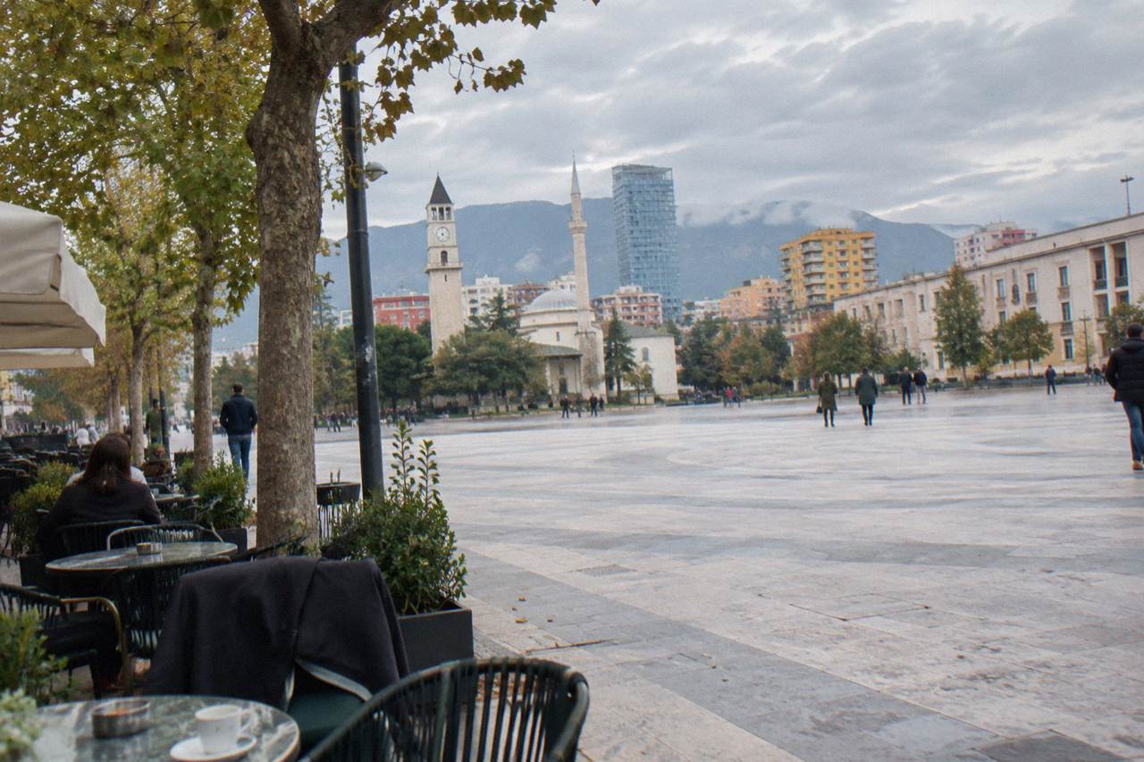 A cafe terrace with small tables and chairs on a tree-lined street in central Tirana