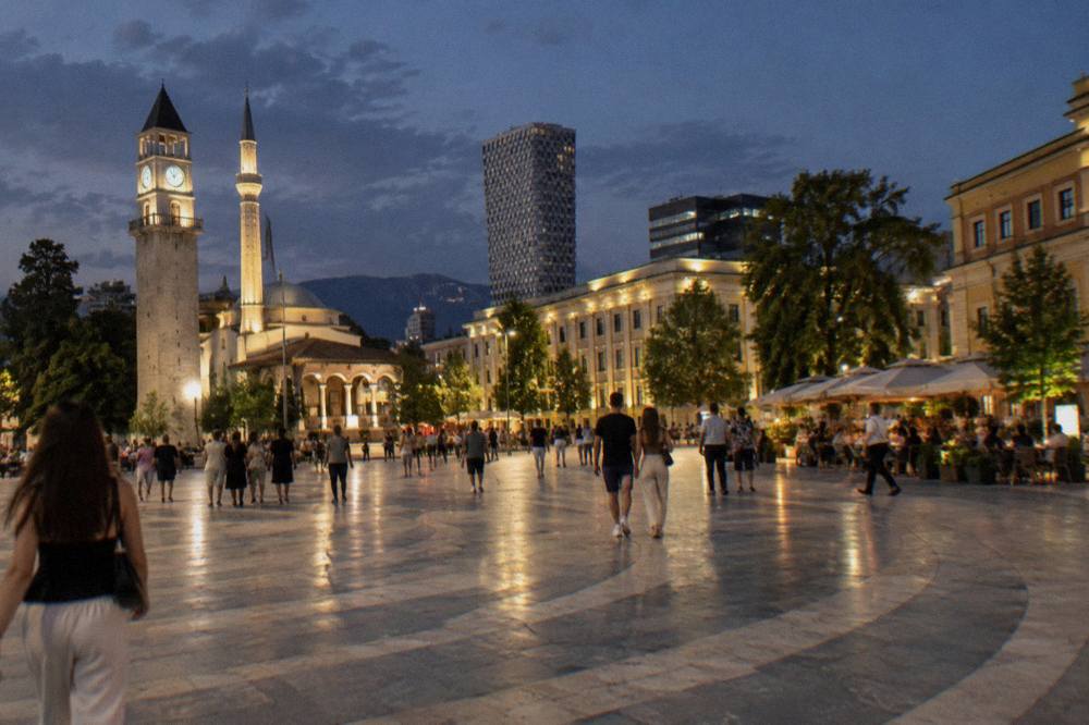 A lit-up square in central Tirana at night with people walking between cafe terraces