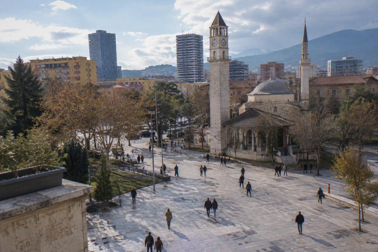 Pedestrians crossing a wide stone-paved plaza in central Tirana with the Clock Tower and mosque minaret behind them