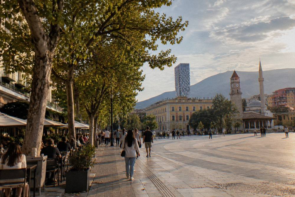 A tree-lined boulevard in Tirana with people strolling under plane trees and cafe tables along the pavement