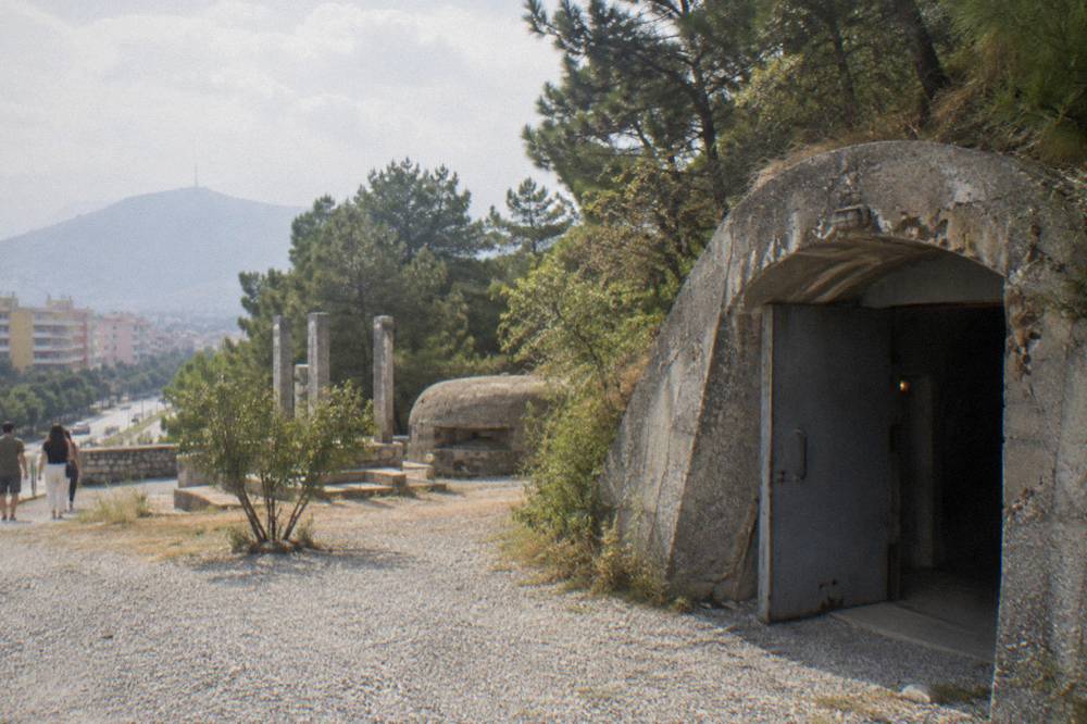The Postbllok memorial with a concrete bunker and stone pillars beside a Tirana boulevard