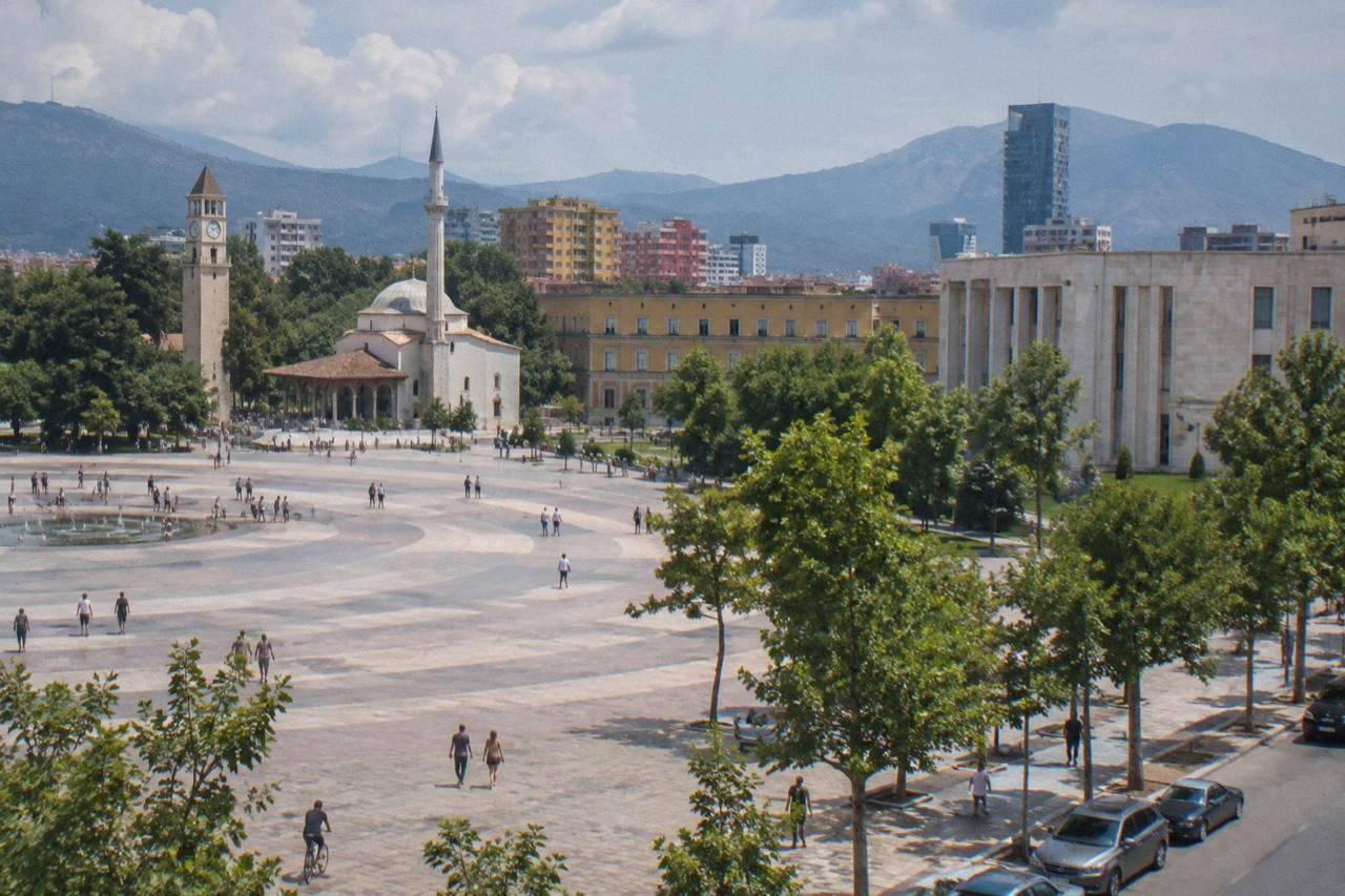 A quiet tree-lined residential street in Tirana with low apartment blocks and parked cars