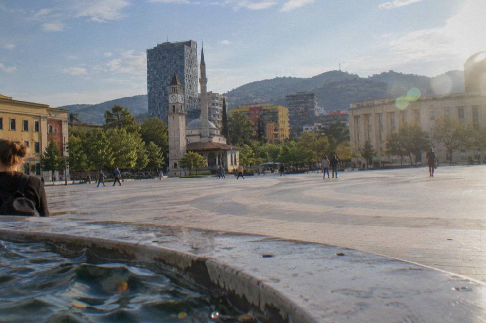 Green hills above Tirana with apartment towers in the foreground under a blue sky