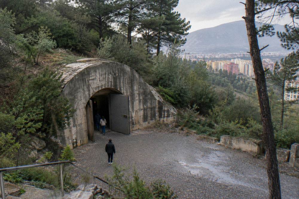 A huge concrete bunker entrance cut into a pine-covered hillside, a plain arched portal with a steel door half open and a gravel forecourt