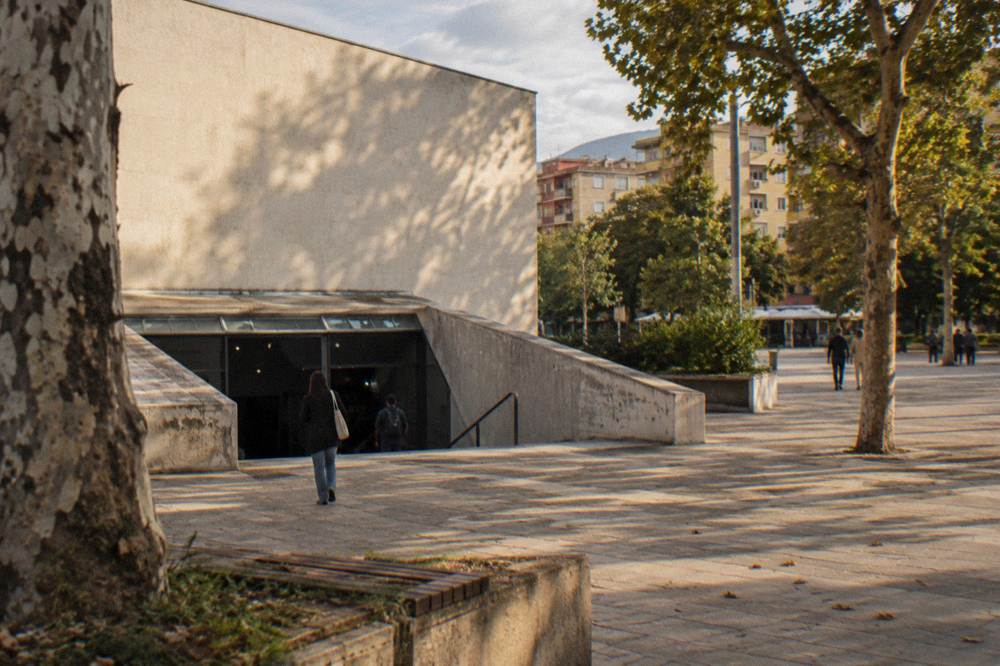 A plain cream concrete-fronted building with a low sloping entrance leading underground beside a small plaza and plane trees