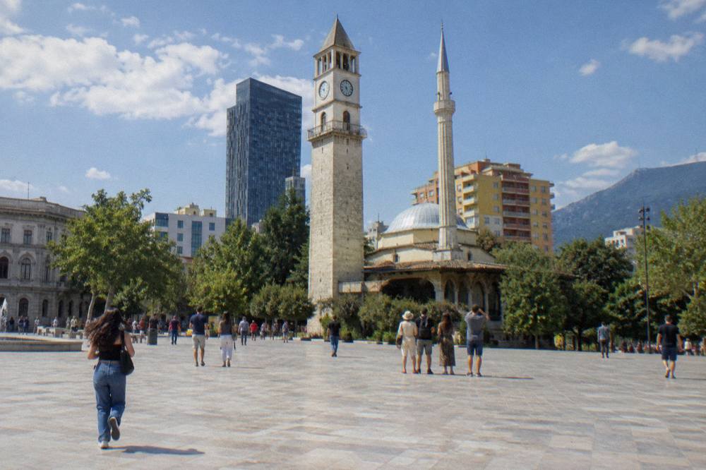 Visitors on Skanderbeg Square looking up at the Clock Tower and the mosque minaret against a blue sky