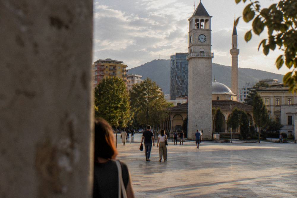 People walking across the wide stone surface of Skanderbeg Square toward the Clock Tower in morning light