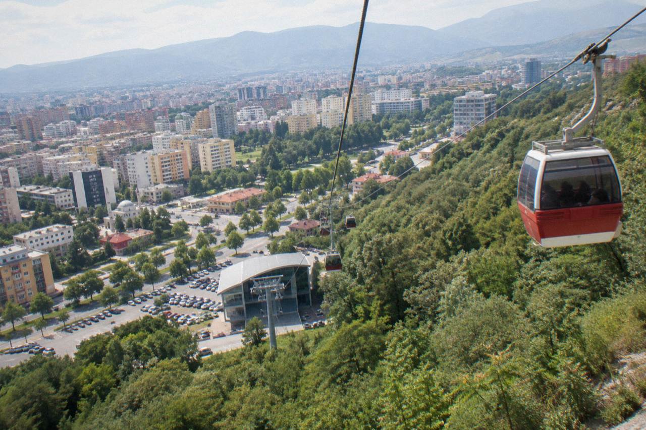 A red and white cable car cabin travelling up a green mountainside above the city of Tirana