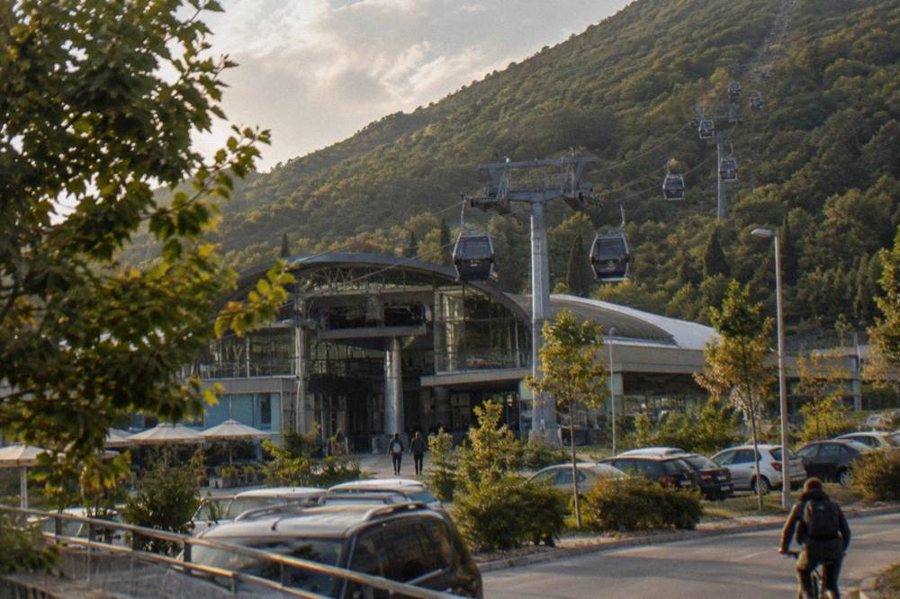 A modern cable-car station of glass and steel at the foot of a long forested mountain ridge with gondolas hanging on cables rising up the slope