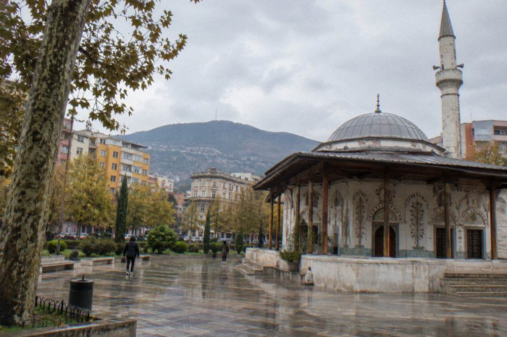 A small early nineteenth-century Ottoman mosque with a low grey dome and a slim pale a slim pale tower beside the dome, a shaded porch with painted floral walls and a quiet paved yard