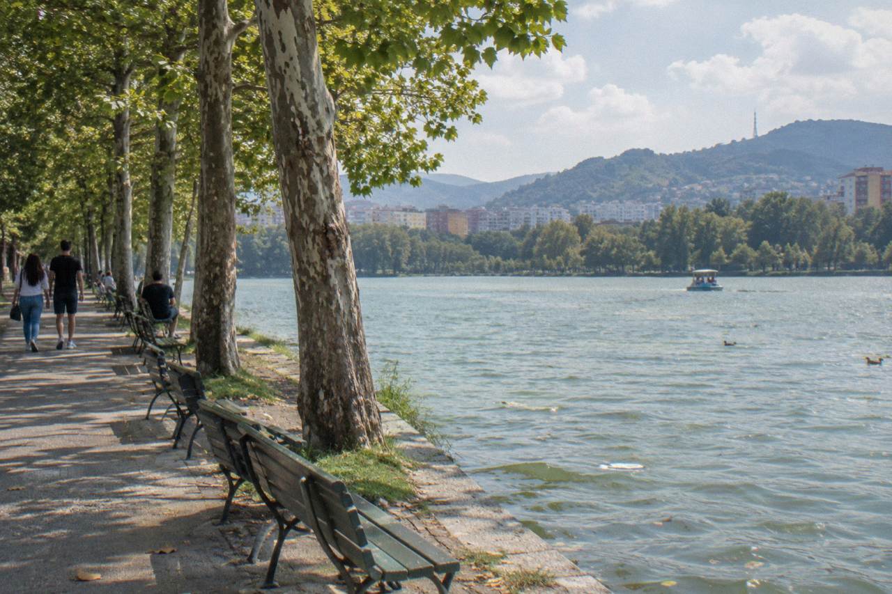 A wide tree-lined path beside the Artificial Lake in the Grand Park of Tirana with hills in the distance