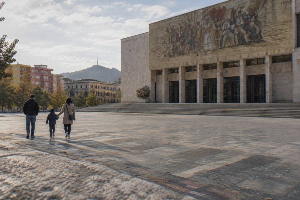 A family walking past the closed museum entrance toward the open square in Tirana