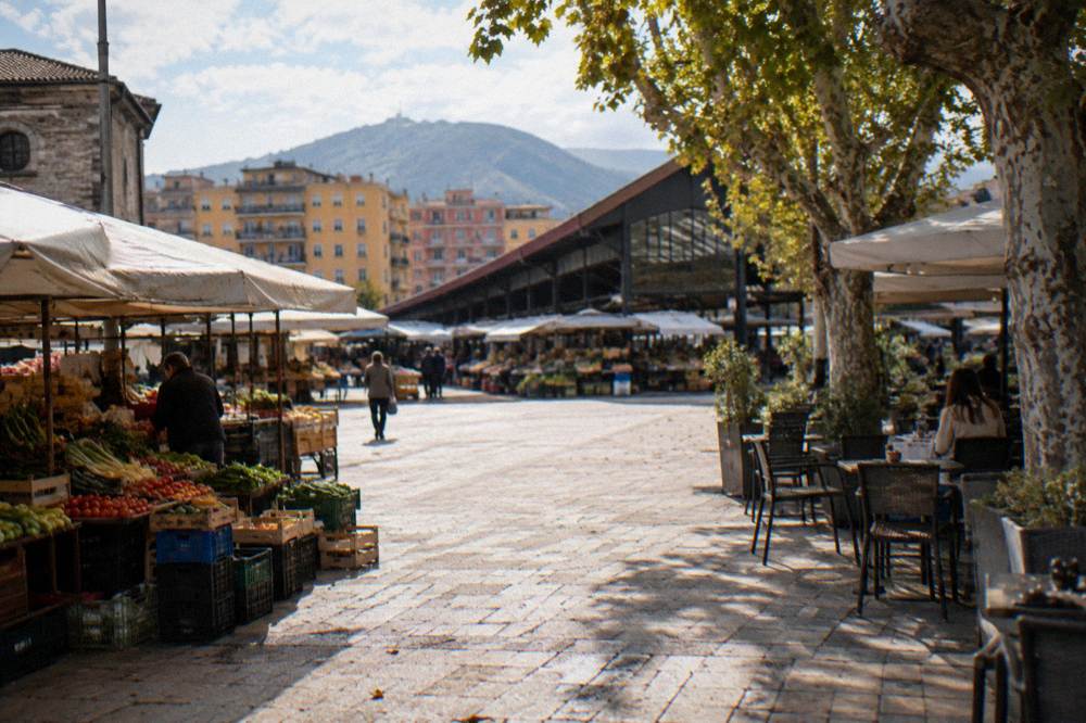 A restored open market square of pale paving with a covered hall, stalls of fresh vegetables and fruit under plain awnings and cafe tables under plane trees