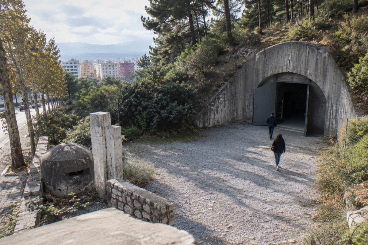 A small roadside memorial of a concrete bunker, stone pillars and a wall fragment beside a leafy boulevard in Tirana