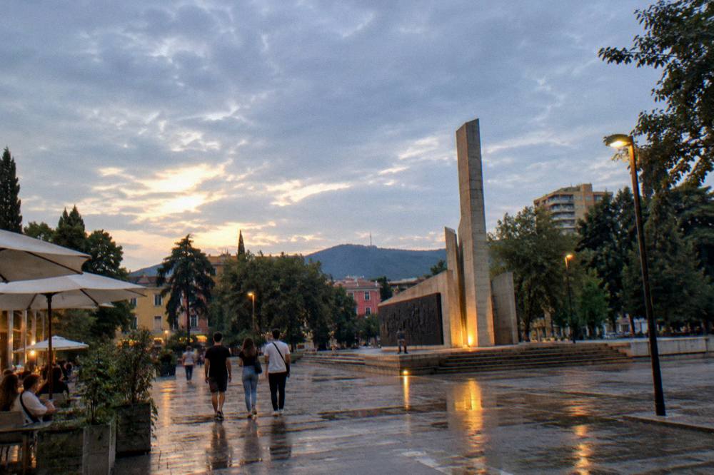 The Postblloku memorial at dusk with street lights and cafe signs glowing in the Blloku quarter behind it