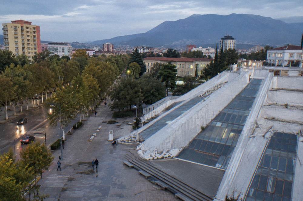 A stepped white concrete pyramid with angled ramps and glass panels at the edge of a wide boulevard of plane trees