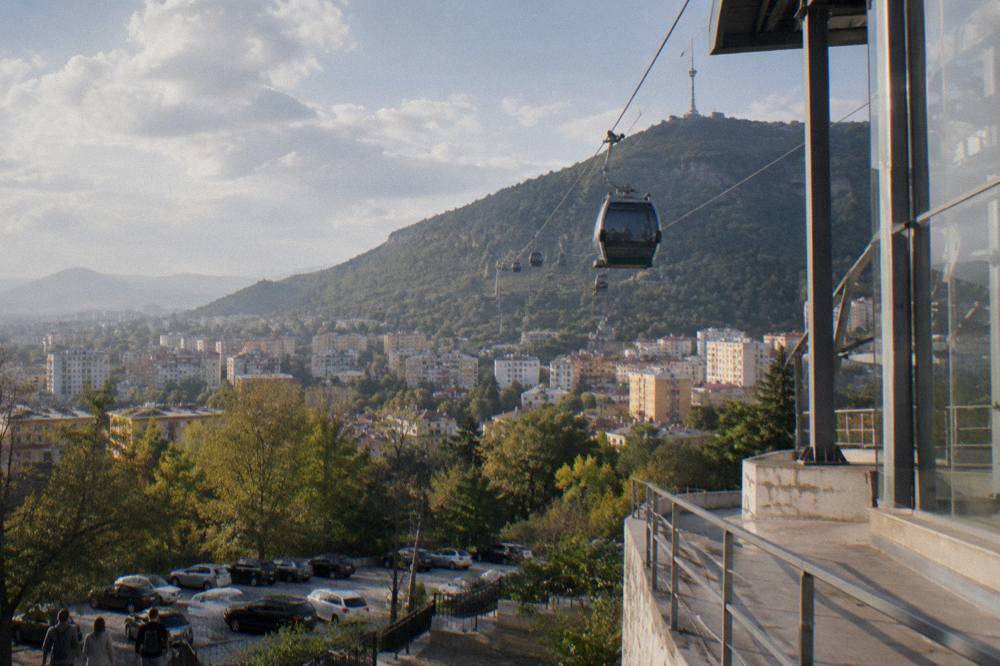 A cable car cabin rising above the city toward the forested ridge of Mount Dajti