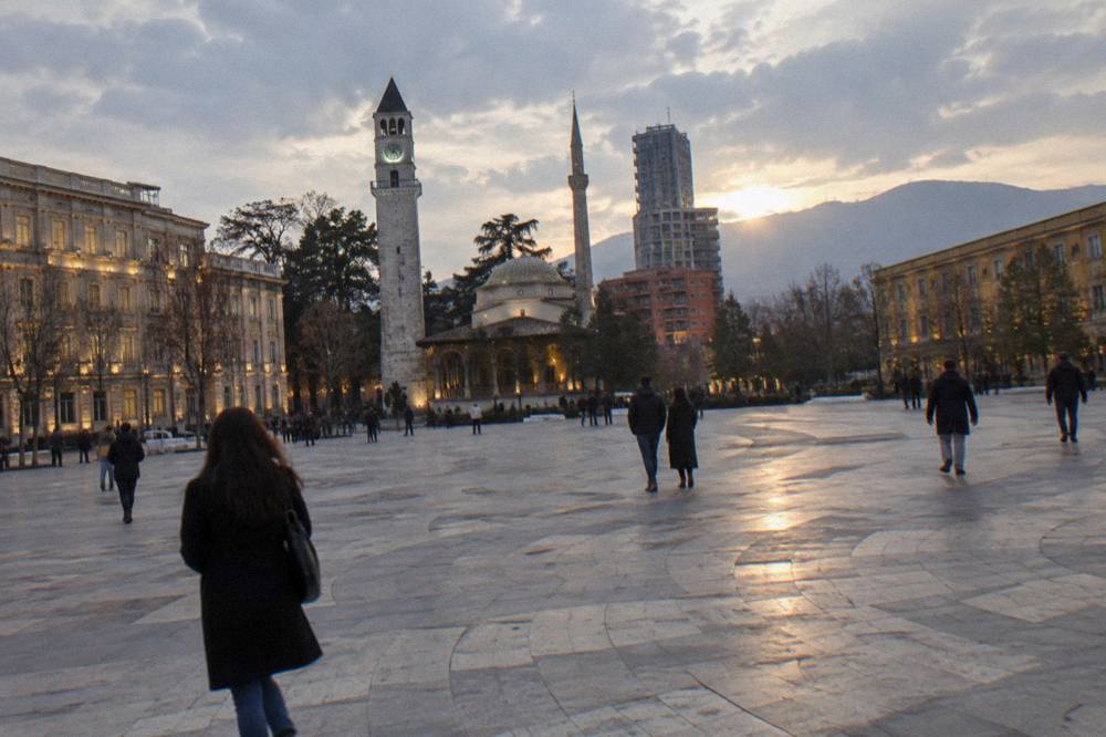People crossing the open stone paving of the square at dusk with lit facades around it